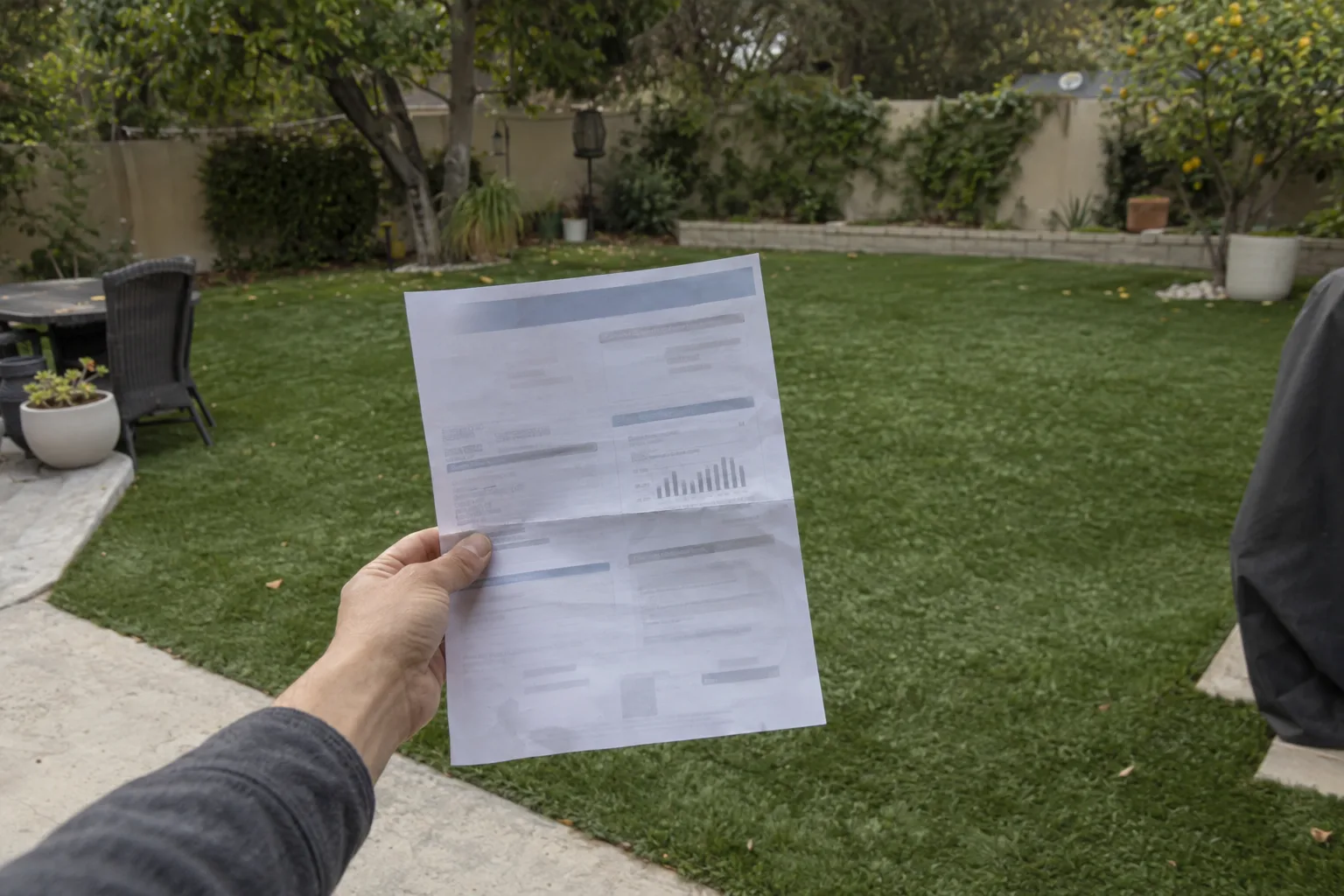 A hand holding a printed utility bill in front of a green artificial turf backyard in Los Angeles, with shallow depth of field and warm afternoon light.