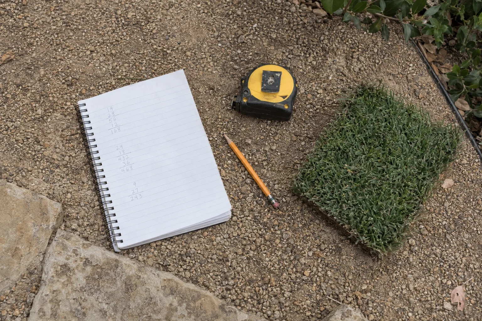 An overhead view of a notepad with handwritten figures, a tape measure, a pencil, and a turf sample swatch arranged on a decomposed-granite surface in bright California daylight.