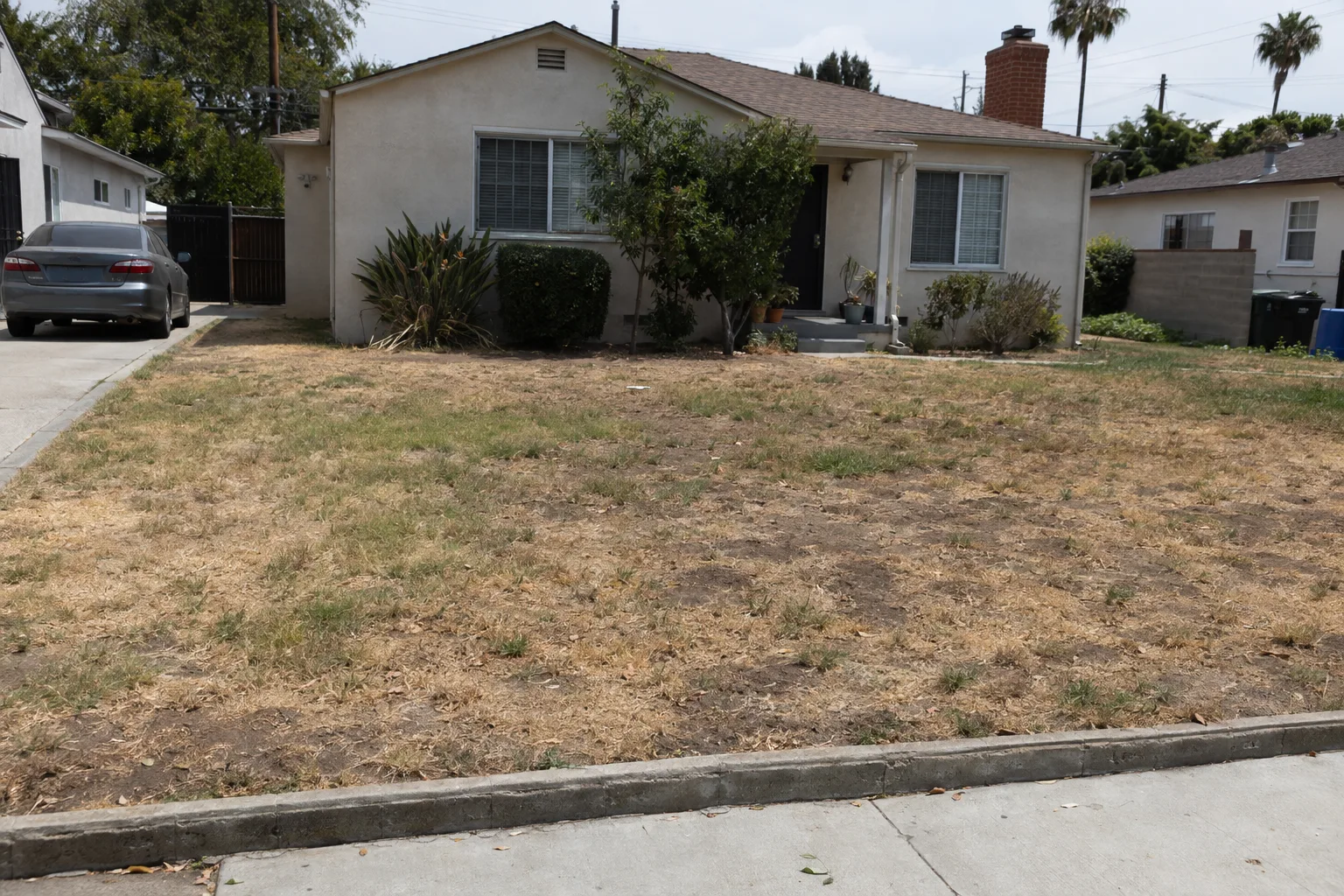 A front yard of a stucco Los Angeles home showing a struggling, patchy lawn with yellowed grass, bare soil patches, and drought damage in bright midday sunlight