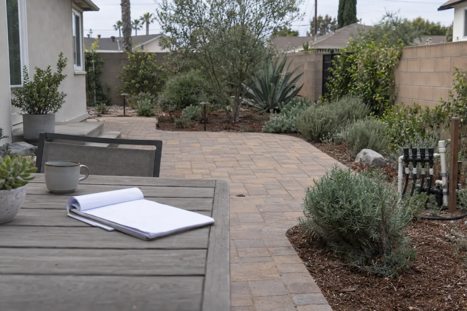 A renovated Los Angeles backyard with stone pavers, a drip-irrigation manifold near a planting bed, drought-tolerant shrubs, and a blank notepad on a patio table under warm afternoon light.
