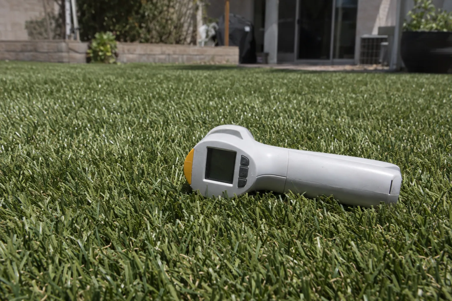 A thermometer probe laid flat on artificial turf under intense midday Southern California sunlight, with the green synthetic blades in sharp focus and a blurred residential yard in the background.