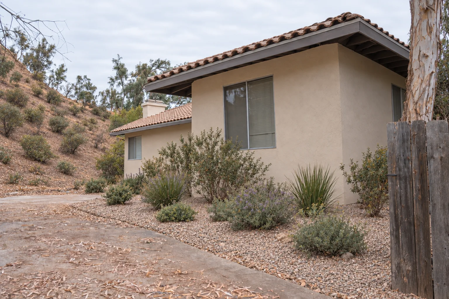 The same hillside stucco house after defensible space clearance work, showing a gravel border against the walls, widely spaced low fire-resistant native plants on the slope, and a roofline clear of overhanging branches