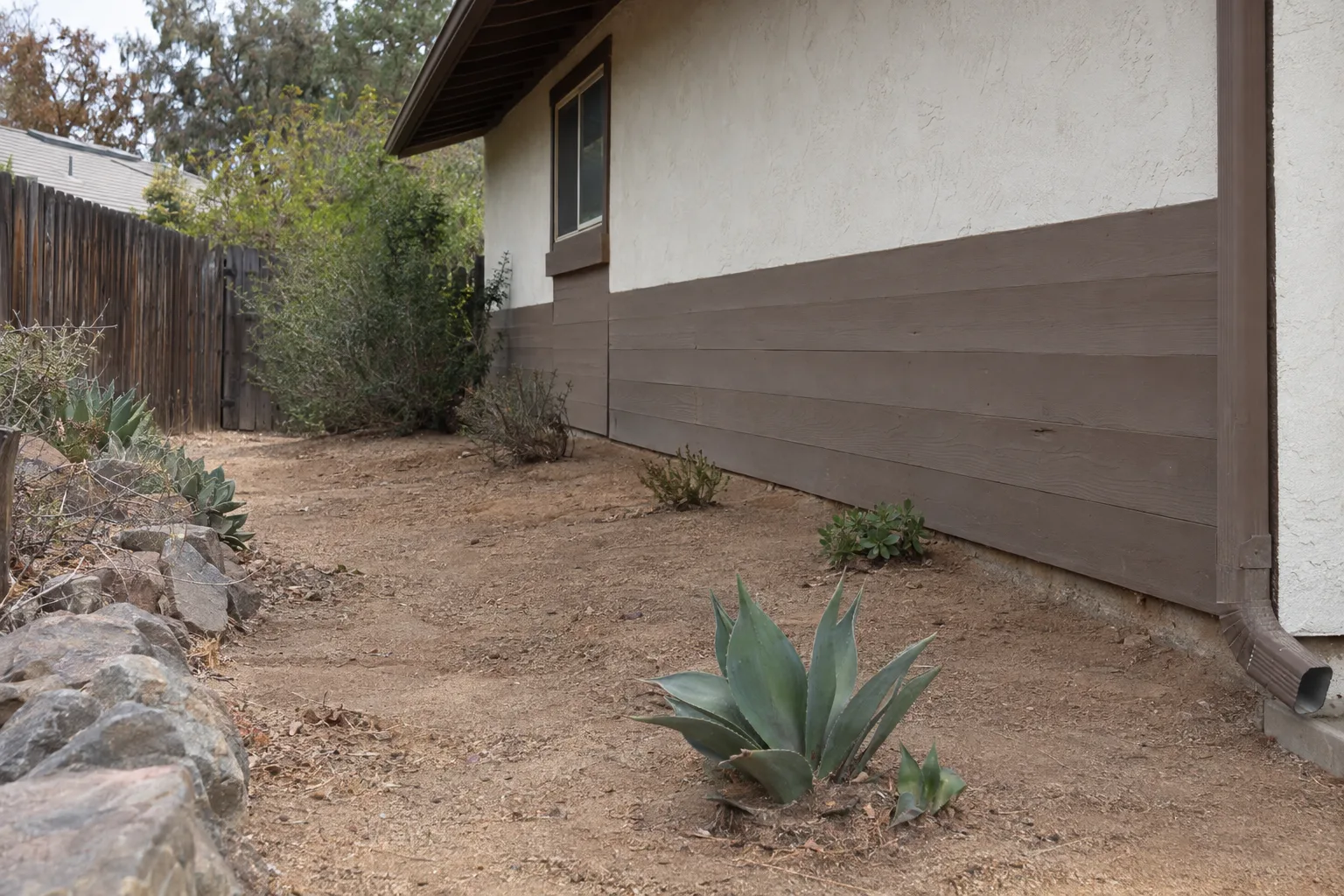 Cleared bare soil and decomposed granite around the foundation and wall of a Southern California home, with removed dry brush and a few remaining low succulents, showing a defensible perimeter