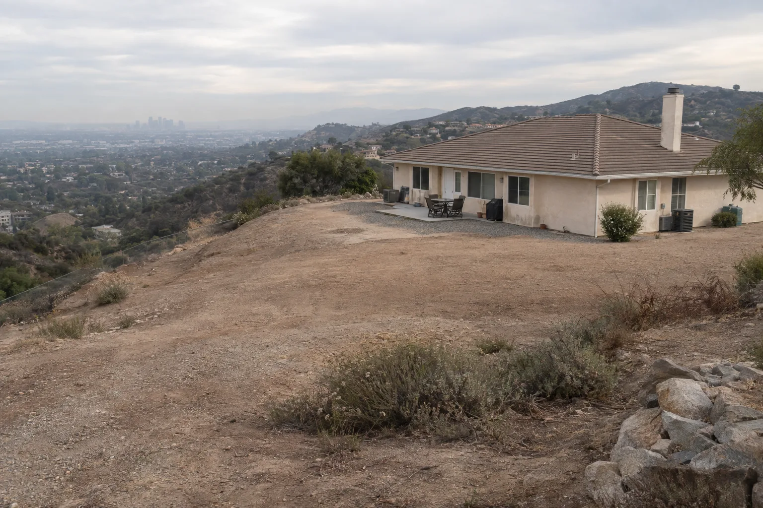 A stucco ranch house on a Southern California hillside surrounded by cleared defensible space, with sparse drought-tolerant plants spaced across the slope and chaparral on the ridgeline behind