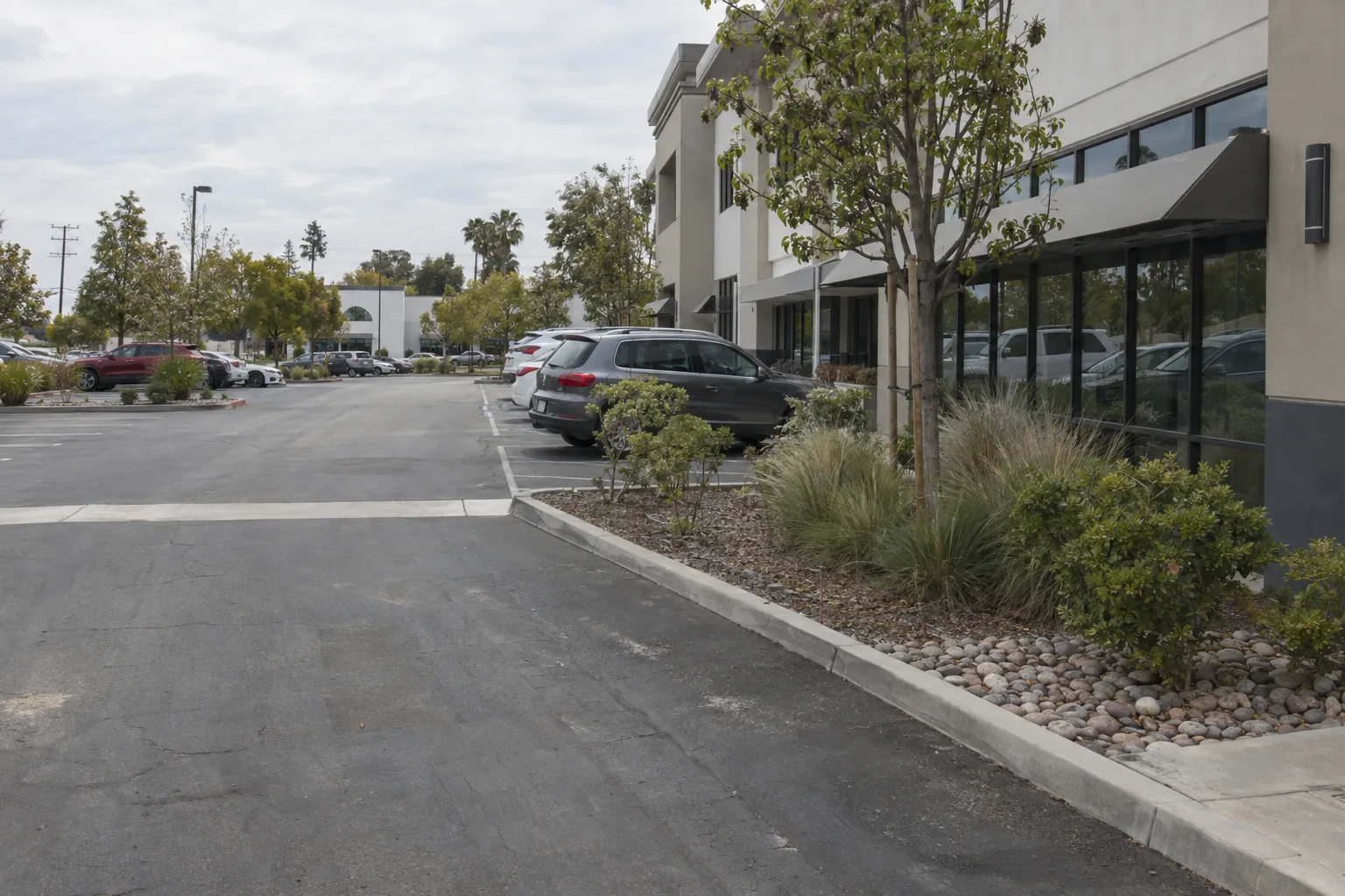 Overhead view of a commercial parking lot with neatly landscaped islands containing ornamental grasses, river rock mulch, and small trees under bright Southern California light