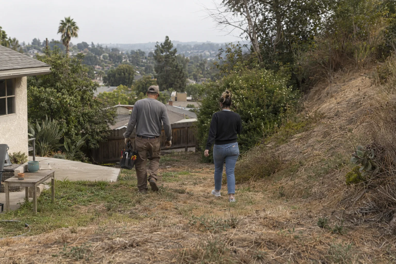 A landscaper and a homeowner walk together across a sloped, partly worn backyard in suburban Los Angeles, seen from behind in warm afternoon light with an eroded hillside visible in the background