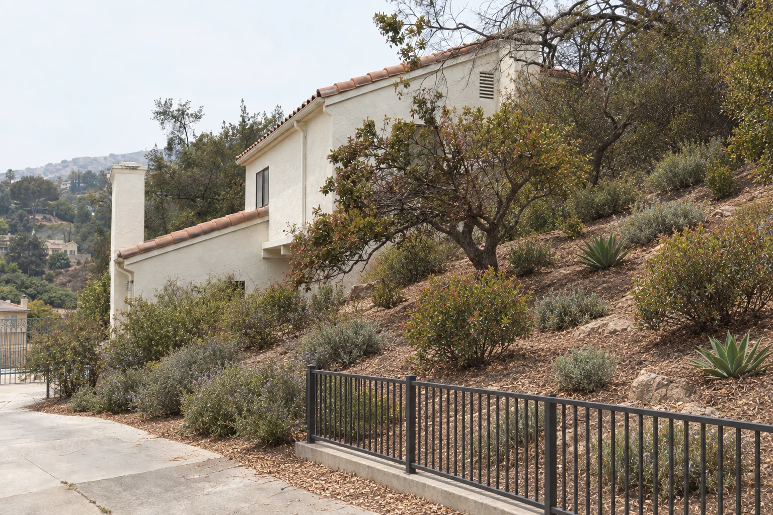 The same Los Angeles hillside stucco house after defensible space work, with a cleared ember-resistant zone at the foundation, widely spaced low fire-resistant native plantings on the slope, and a metal fence replacing the original wooden one