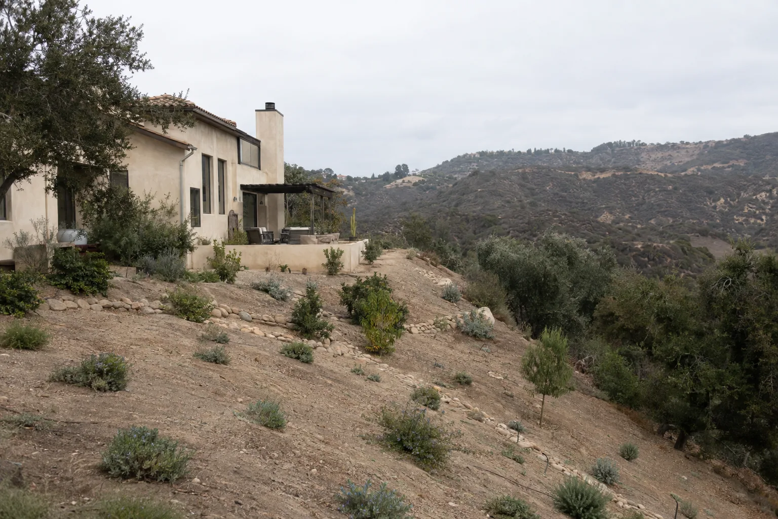 A hillside house in the Santa Monica Mountains with cleared defensible space on the surrounding slope, sparse low-growing groundcover visible in the foreground, chaparral ridgeline in the background under a warm golden-hour sky
