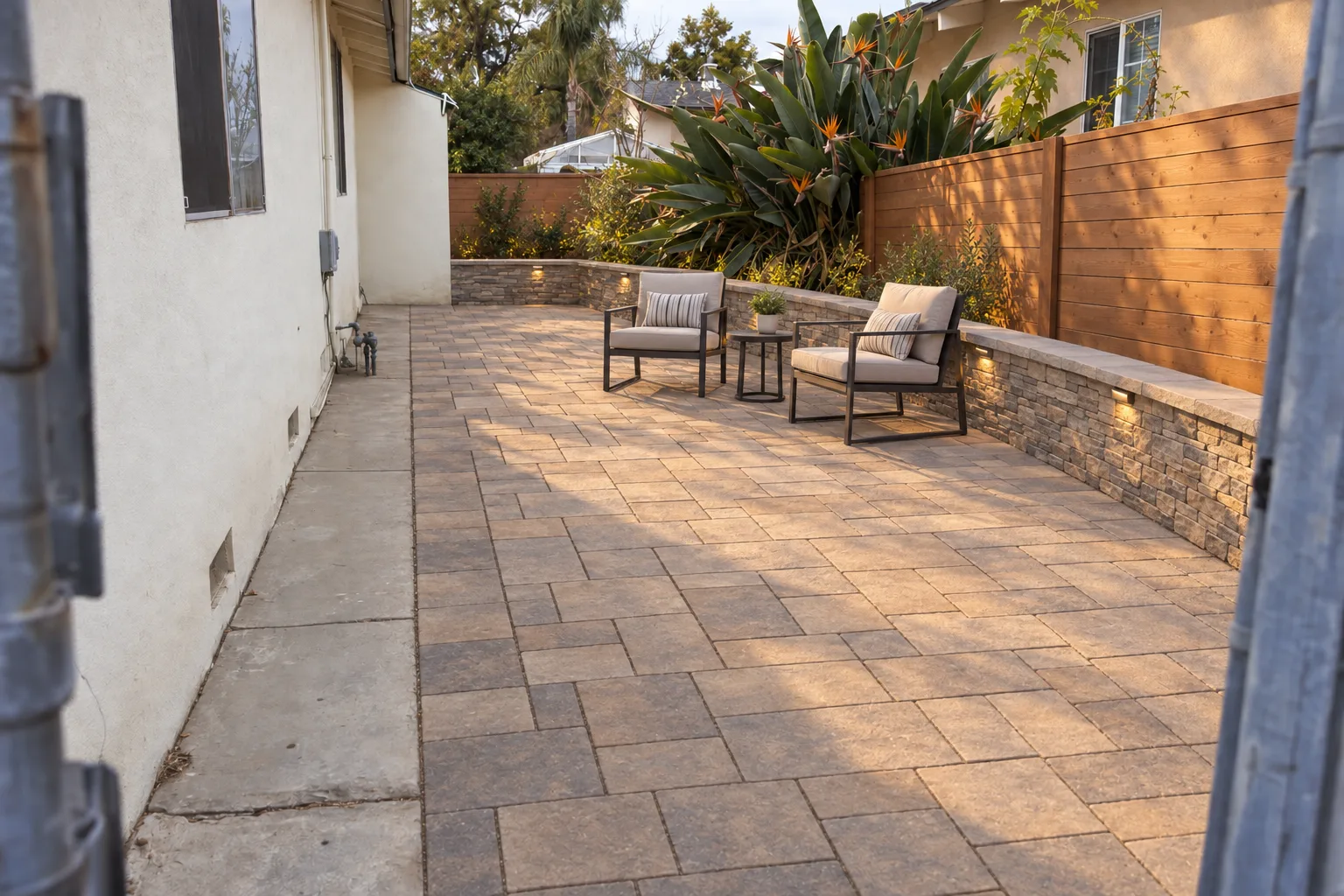 A finished paver patio beside a stucco ranch house in Los Angeles, bordered by a low stacked-stone seat wall and a wood fence, with two outdoor chairs on the pavers in warm late-afternoon light