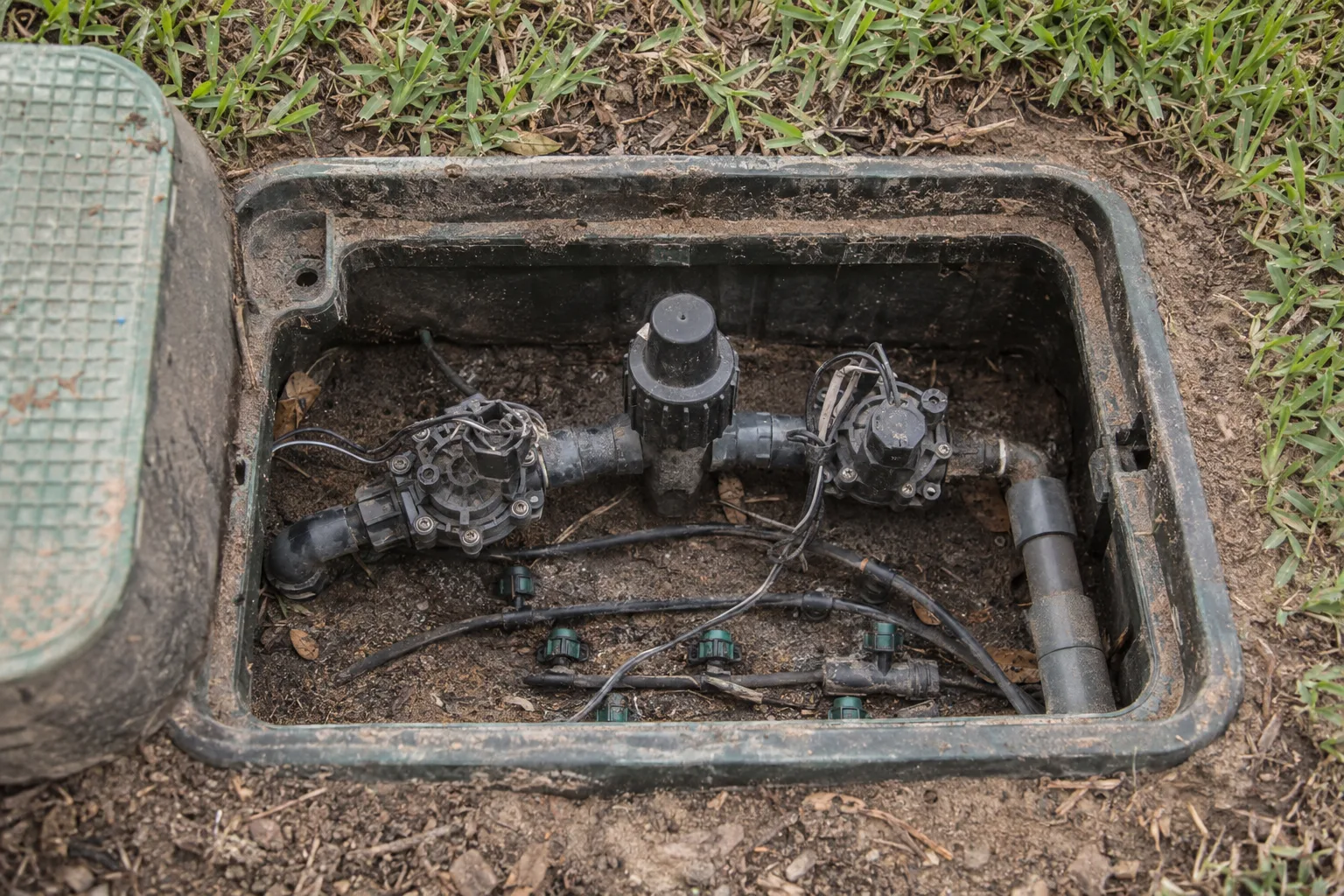 An open residential irrigation valve box embedded in a lawn, showing drip emitters and a pressure regulator inside, surrounded by sandy Southern California soil