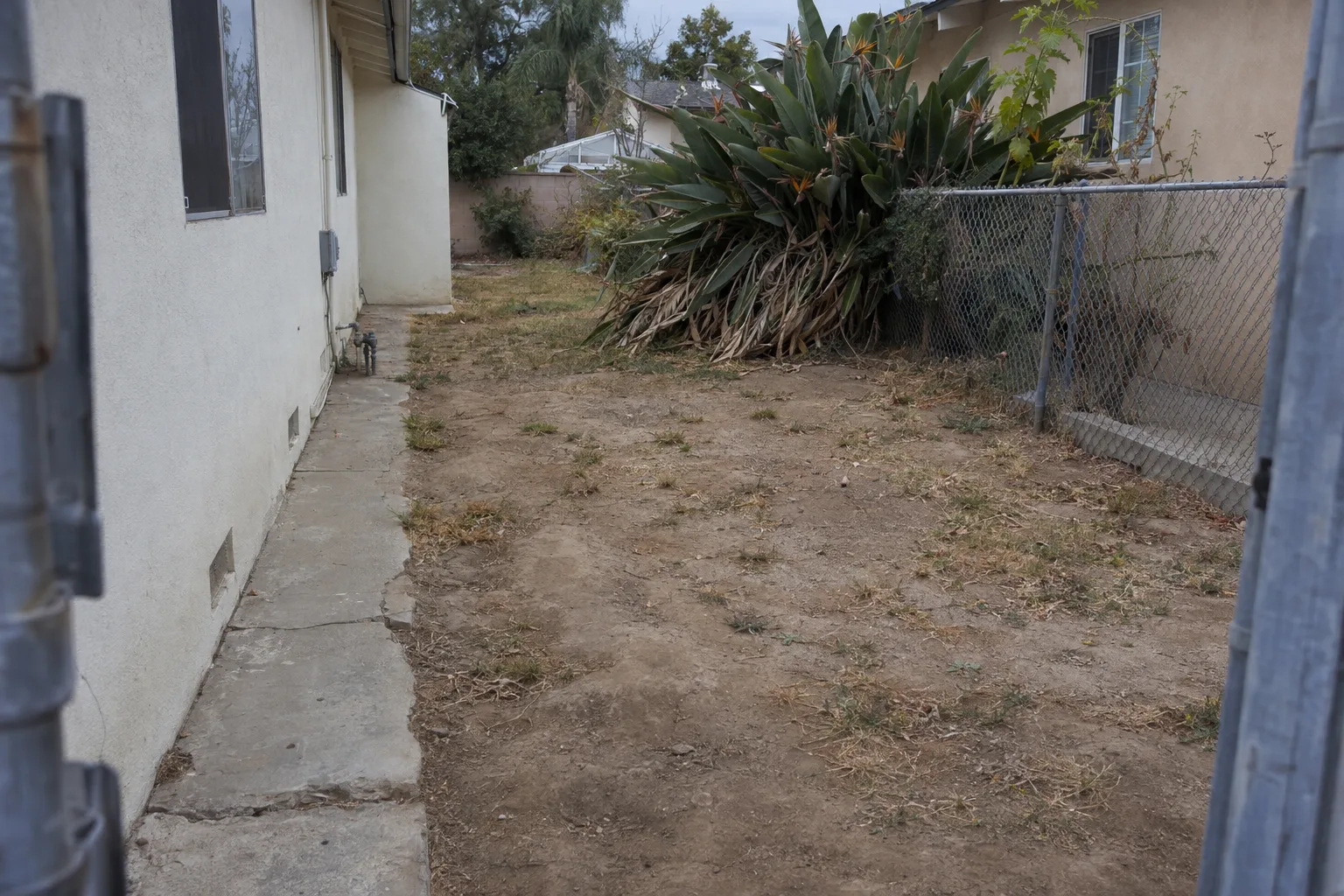 A bare, compacted-dirt side yard beside a stucco ranch house in Los Angeles, with sparse dry grass, a cracked concrete strip, and overgrown plants, showing an unused outdoor space before any hardscape work