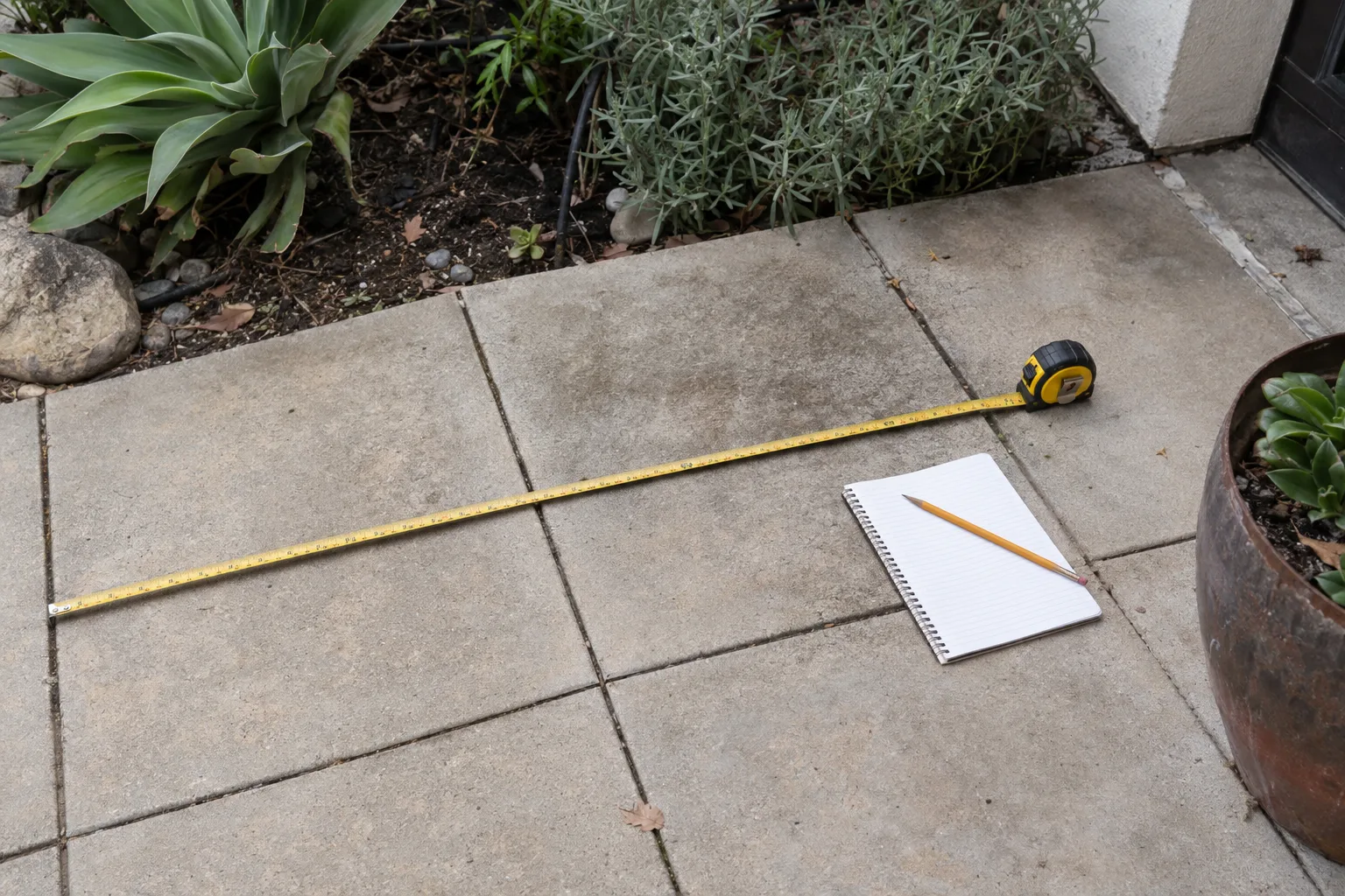 A measuring tape and notepad on a concrete paver patio surface with a garden bed in the background, shot from overhead in natural daylight