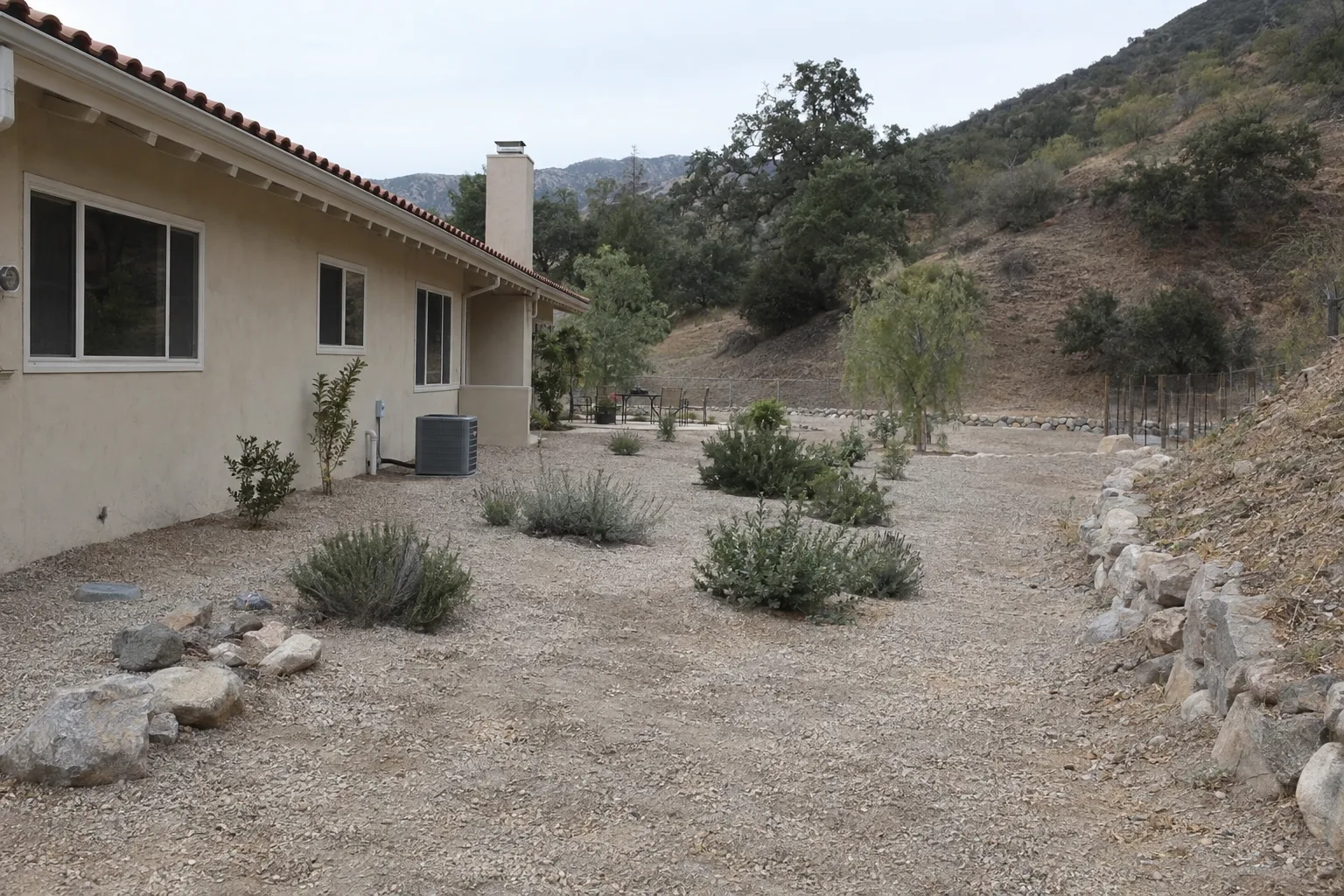 A fire-safe residential yard in the LA foothills with cleared defensible space, gravel ground cover, widely spaced low plants, and no combustible material near the stucco home foundation