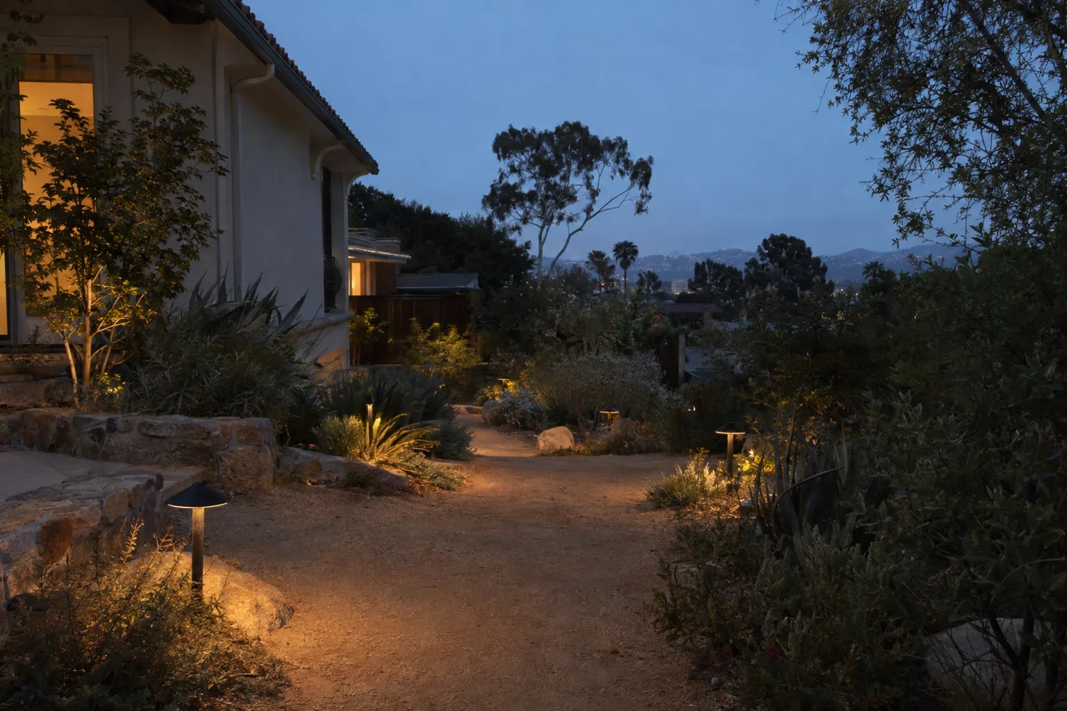 Low-voltage path lights glowing amber along a decomposed-granite garden walkway at dusk, with native plantings lit on either side under a deep blue sky