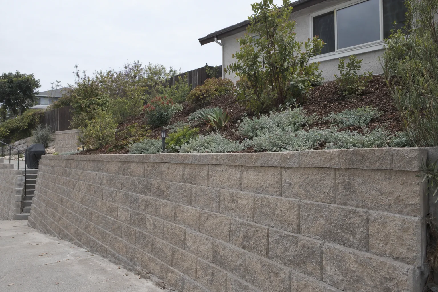 A finished concrete-block retaining wall on an LA residential slope with a planted terraced bed of groundcover and ornamental shrubs above it