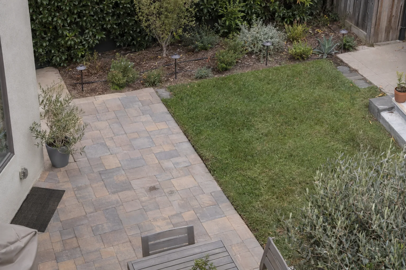 An overhead view of a Los Angeles residential yard showing a small lawn area, a paver patio, a drip-irrigated native planting bed, and low path lighting along a garden border.