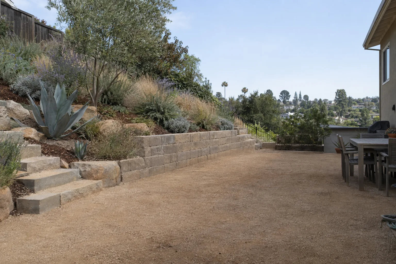 A completed Los Angeles backyard featuring a decomposed-granite patio, a stacked-block retaining wall on a gentle slope, and native drought-tolerant plantings in afternoon sunlight