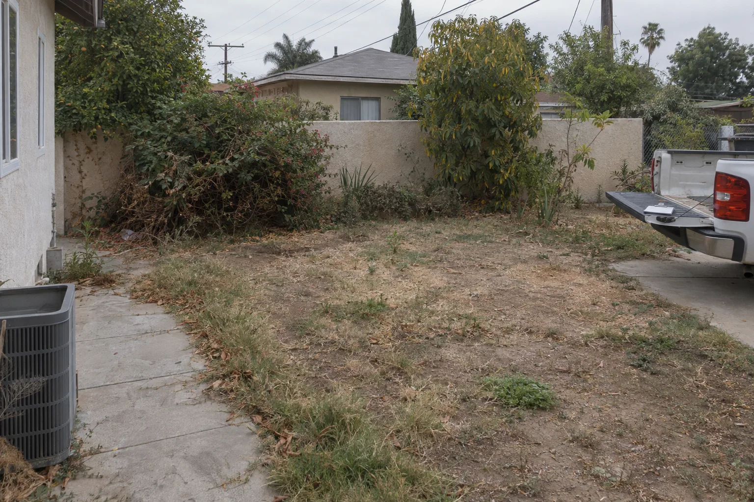 A neglected Los Angeles backyard with dead grass, bare dirt patches, overgrown shrubs, and a stucco wall, photographed before landscaping work begins, with an unbranded clipboard resting on a truck tailgate in the background.