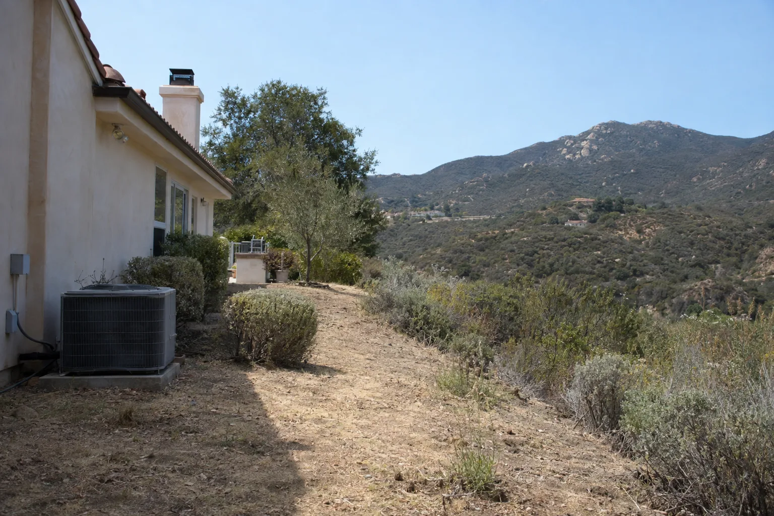 A hillside Los Angeles home with a cleared Zone 0 and Zone 1 defensible-space buffer, short dry grass close to the stucco structure transitioning to sparse native shrubs, chaparral ridge visible behind