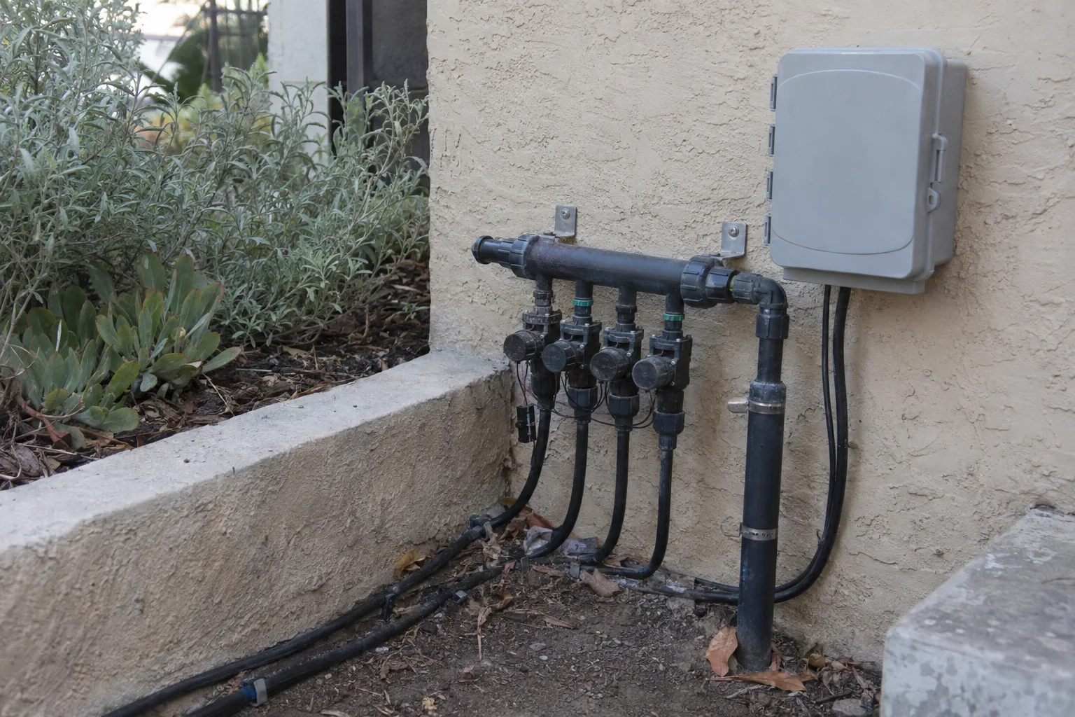A drip-irrigation manifold and smart controller mounted on a stucco exterior wall beside a raised planter of native plants at a Los Angeles residence, photographed in soft morning light