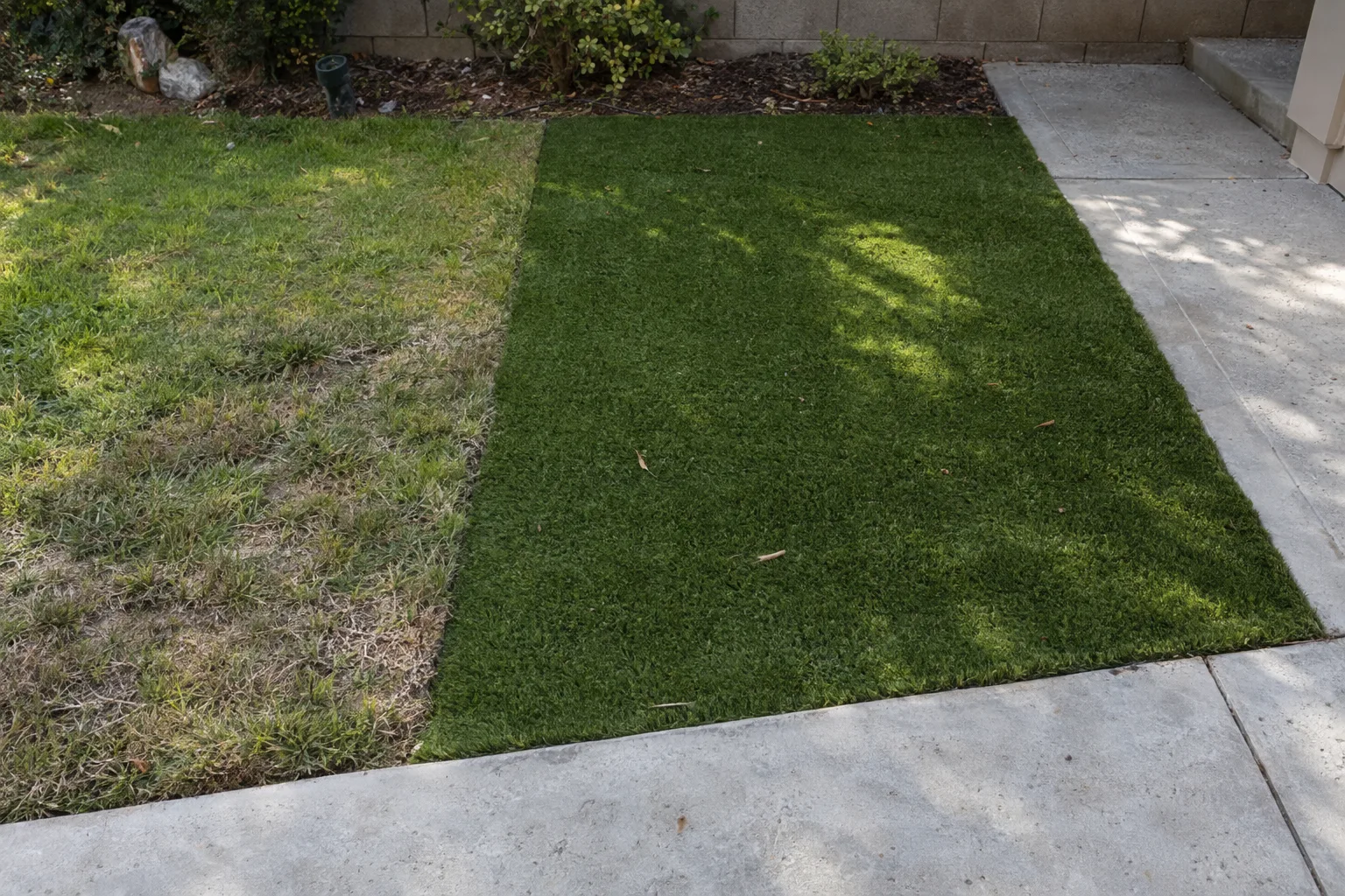 A close overhead view of freshly installed artificial turf meeting a concrete patio edge in a suburban Los Angeles backyard, with a strip of natural grass alongside in afternoon light