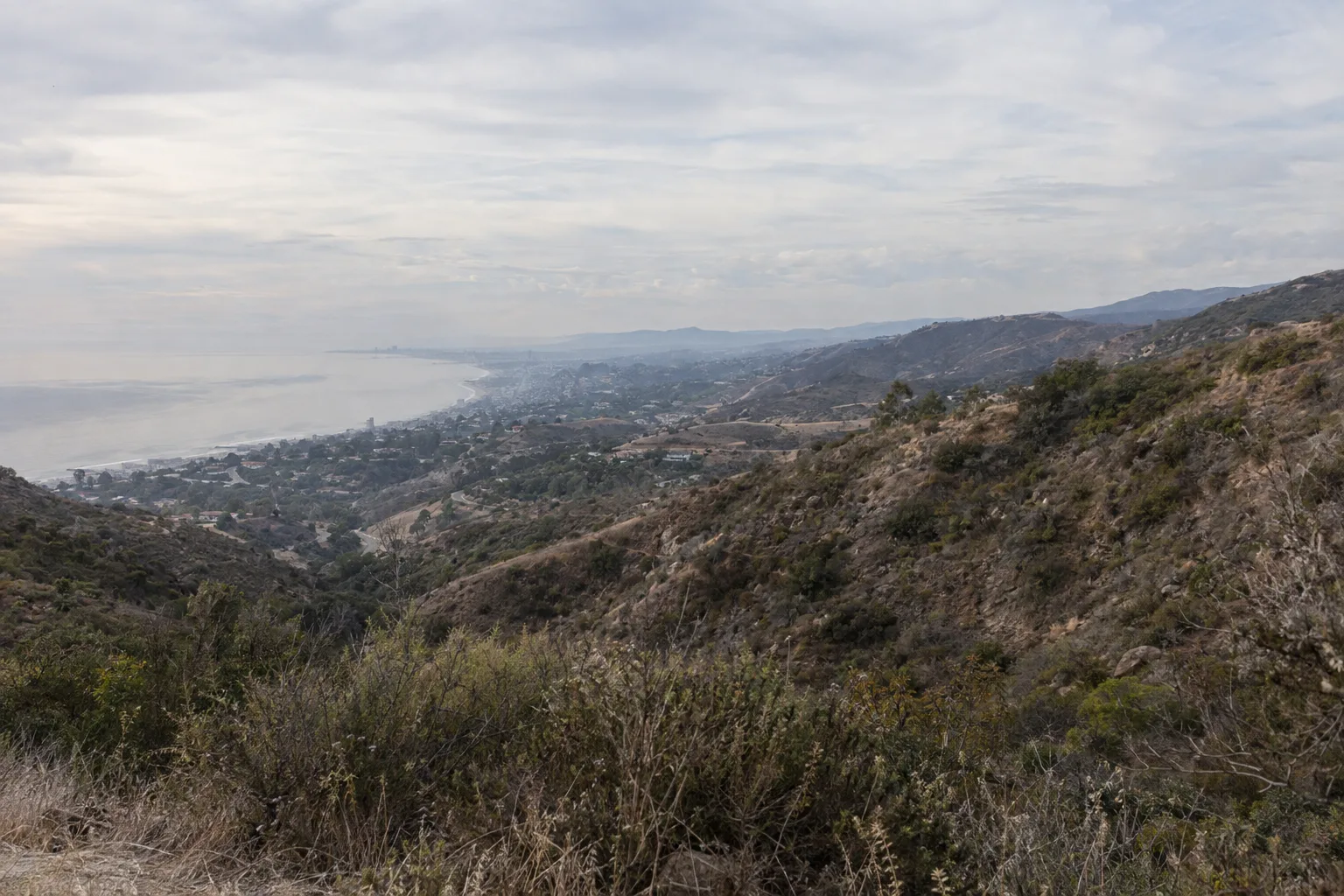 Panoramic view from a Santa Monica Mountains ridgeline showing the Los Angeles coastal basin with marine layer on the left and dry, sun-exposed inland chaparral hills on the right at golden hour