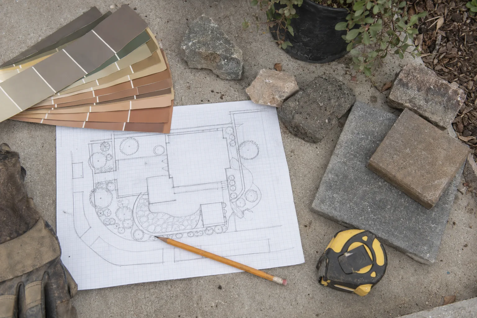 A flat-lay of hardscape planning materials including a pencil site sketch on graph paper, paver samples, stone chips, and a tape measure on a concrete surface
