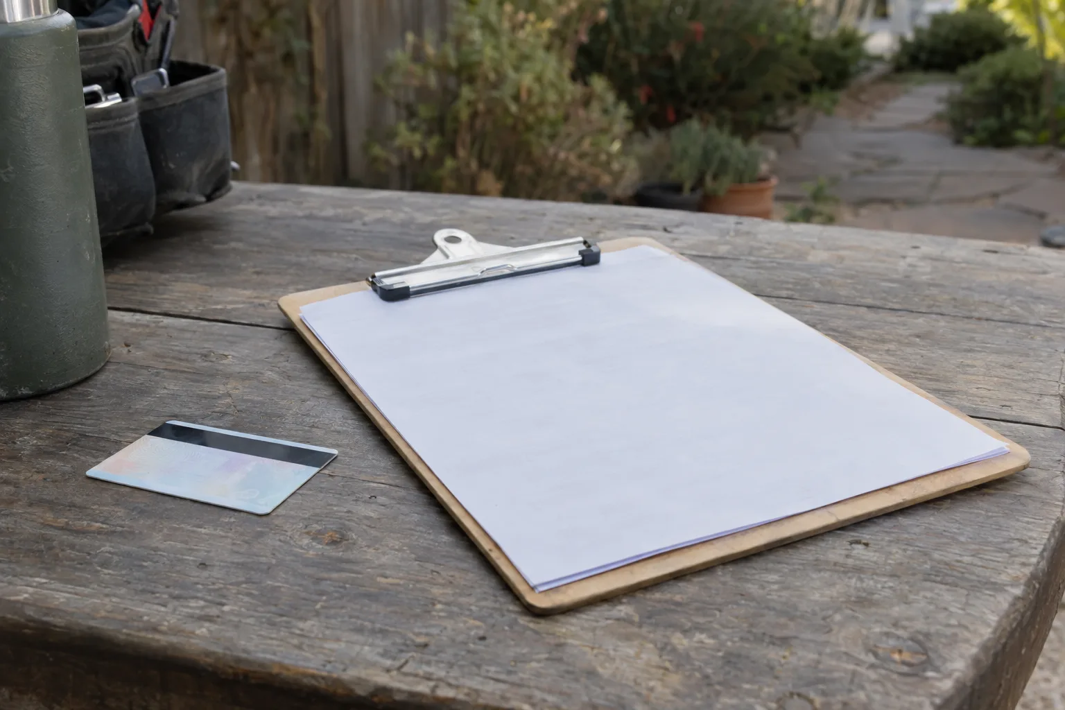 A contractor licence card and insurance certificate on a clipboard resting on a wooden outdoor table, face-down, with a blurred garden in the background