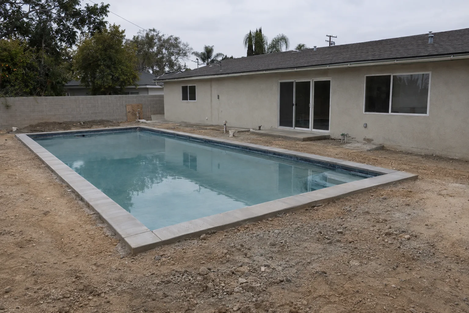 A newly installed in-ground pool surrounded by bare compacted dirt in a Los Angeles backyard, with raw coping edges, no plants or decking, and a plain stucco ranch house visible in the background.