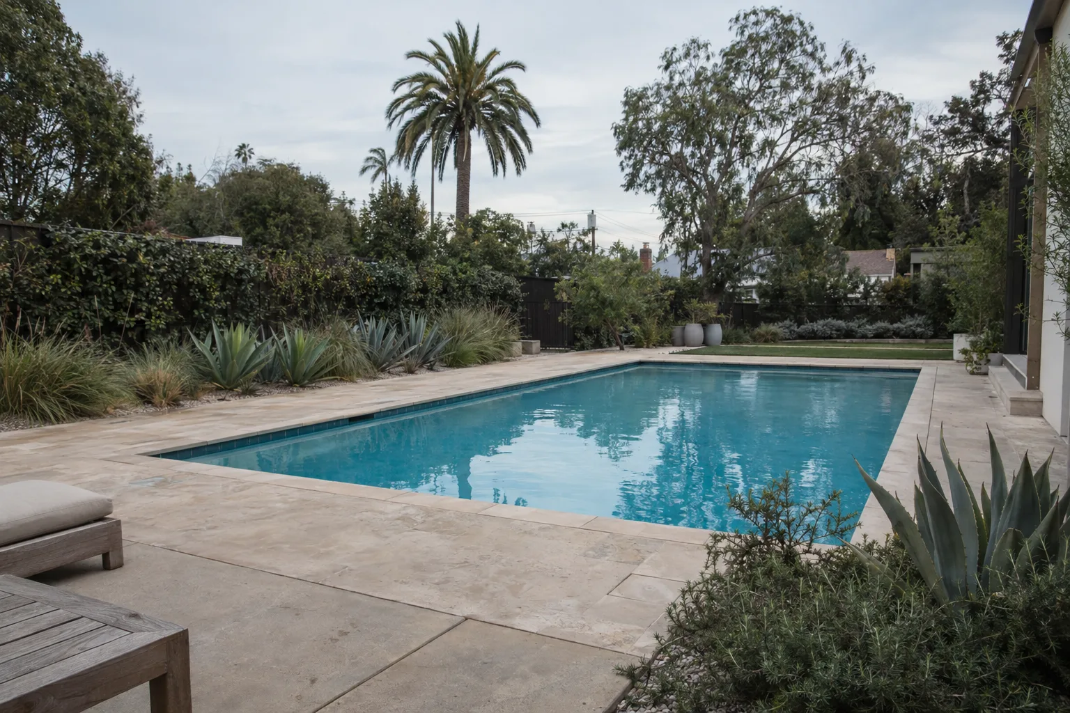 A finished backyard pool with travertine decking, agave and ornamental grass plantings along the edges, and a palm tree against a bright Los Angeles sky