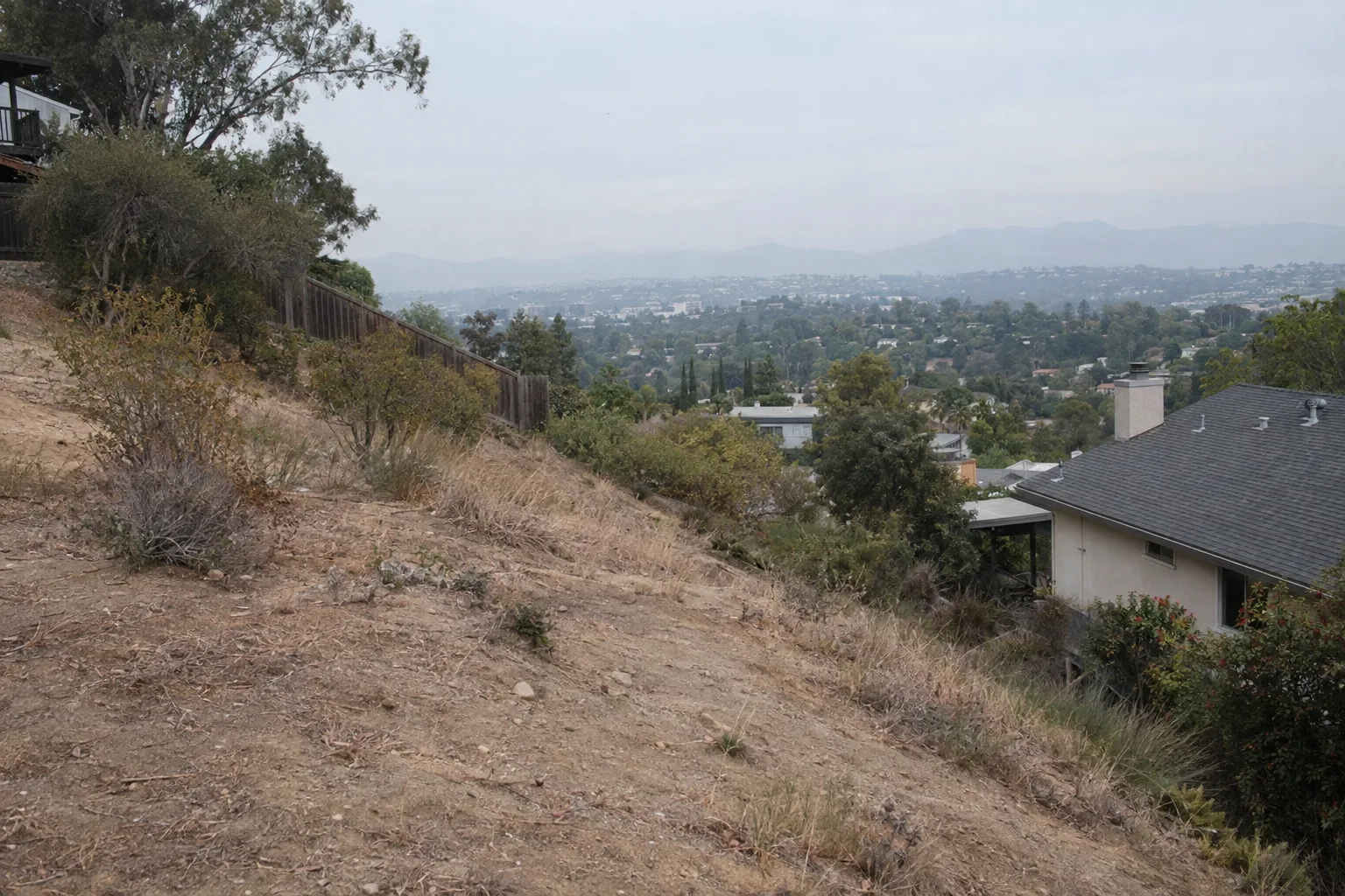 A dry, sloped hillside residential lot in Los Angeles with sparse vegetation and compacted clay soil in late afternoon light