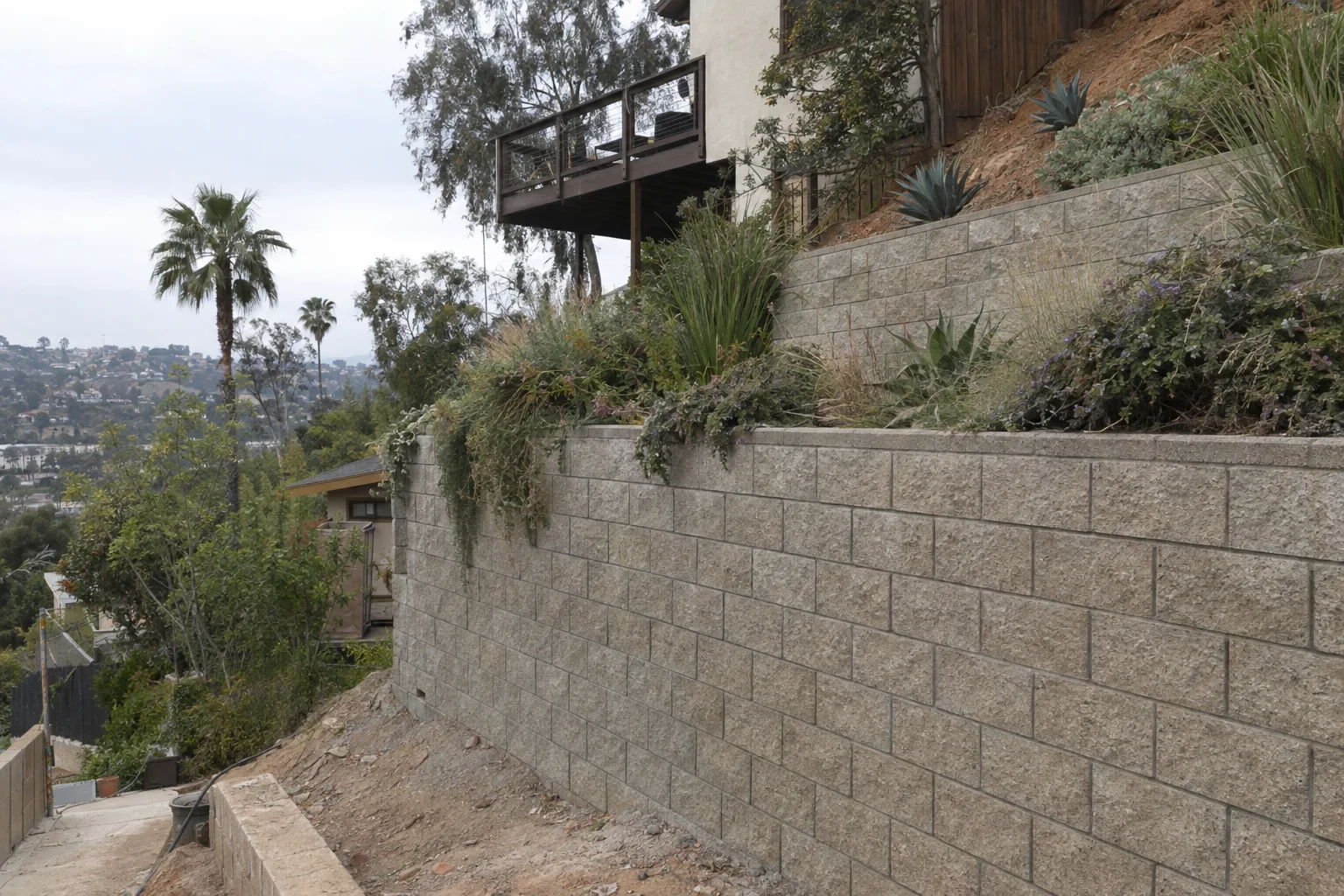 A completed multi-tiered concrete block retaining wall terracing a steep hillside lot in Los Angeles, with drought-tolerant plants along the upper tier and palm trees visible against the hazy sky in the background.