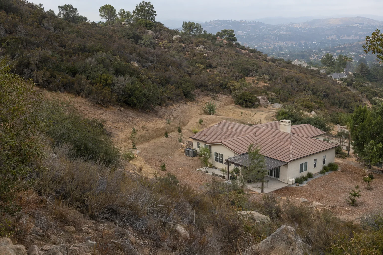 An aerial view of a stucco home on a dry Southern California hillside with a cleared defensible-space perimeter surrounding it and dense chaparral brush beginning further up the slope
