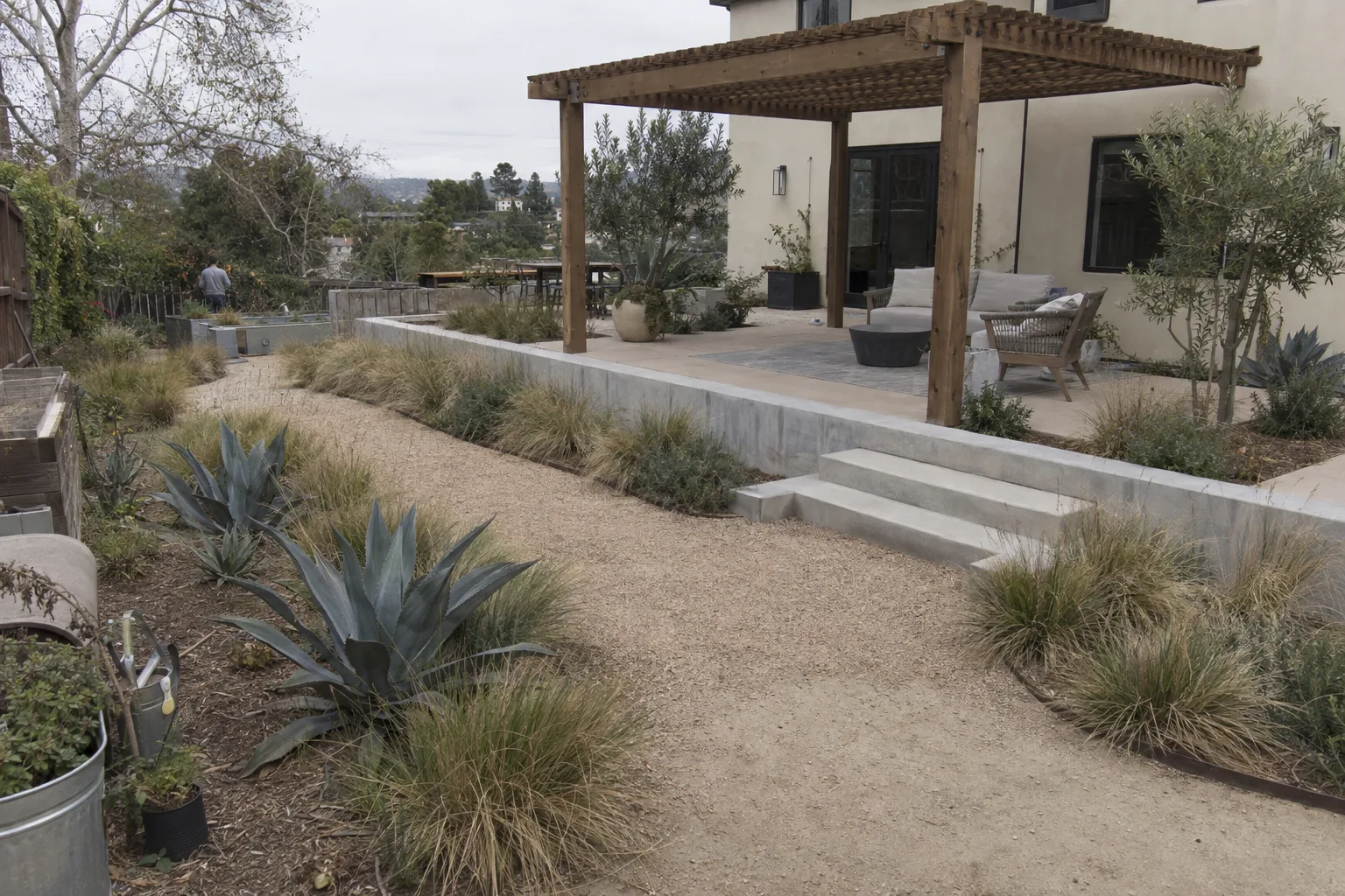 A fully designed Los Angeles backyard at golden hour featuring decomposed-granite paths, agave and ornamental grasses, a concrete step, and a wooden pergola over an outdoor seating area