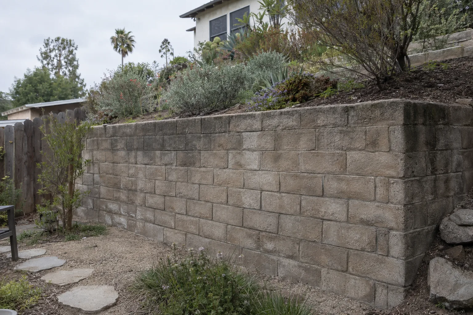 A stacked-concrete-block retaining wall about four feet high holding back a terraced hillside garden planted with drought-tolerant native shrubs at a Los Angeles residential property, photographed in diffused overcast light