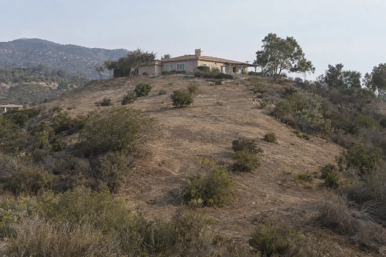 A Southern California hillside cleared for defensible space with dry scrub removed in a wide perimeter around a stucco home at the crest, leaving only widely spaced fire-resistant native shrubs in late-afternoon hazy sunlight
