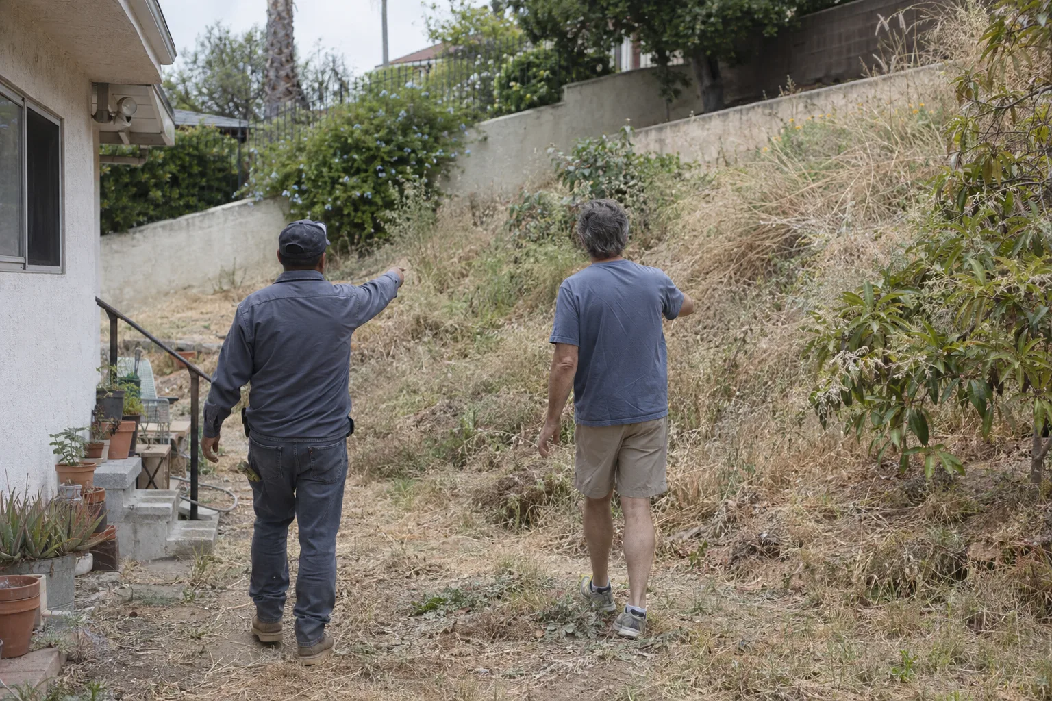 Two people seen from behind walking a dry residential lot in Los Angeles, one in work attire gesturing toward an overgrown slope, stucco wall and dry hills in the background