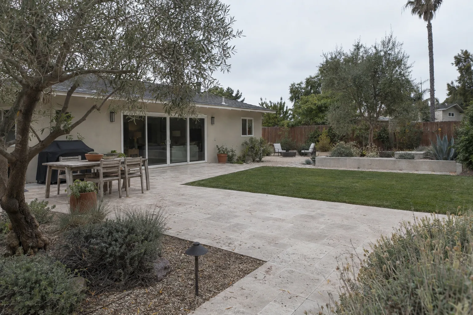 A finished Los Angeles backyard featuring a travertine paver patio, drought-tolerant plantings, a trimmed olive tree, and a manicured lawn panel bathed in late-afternoon golden light