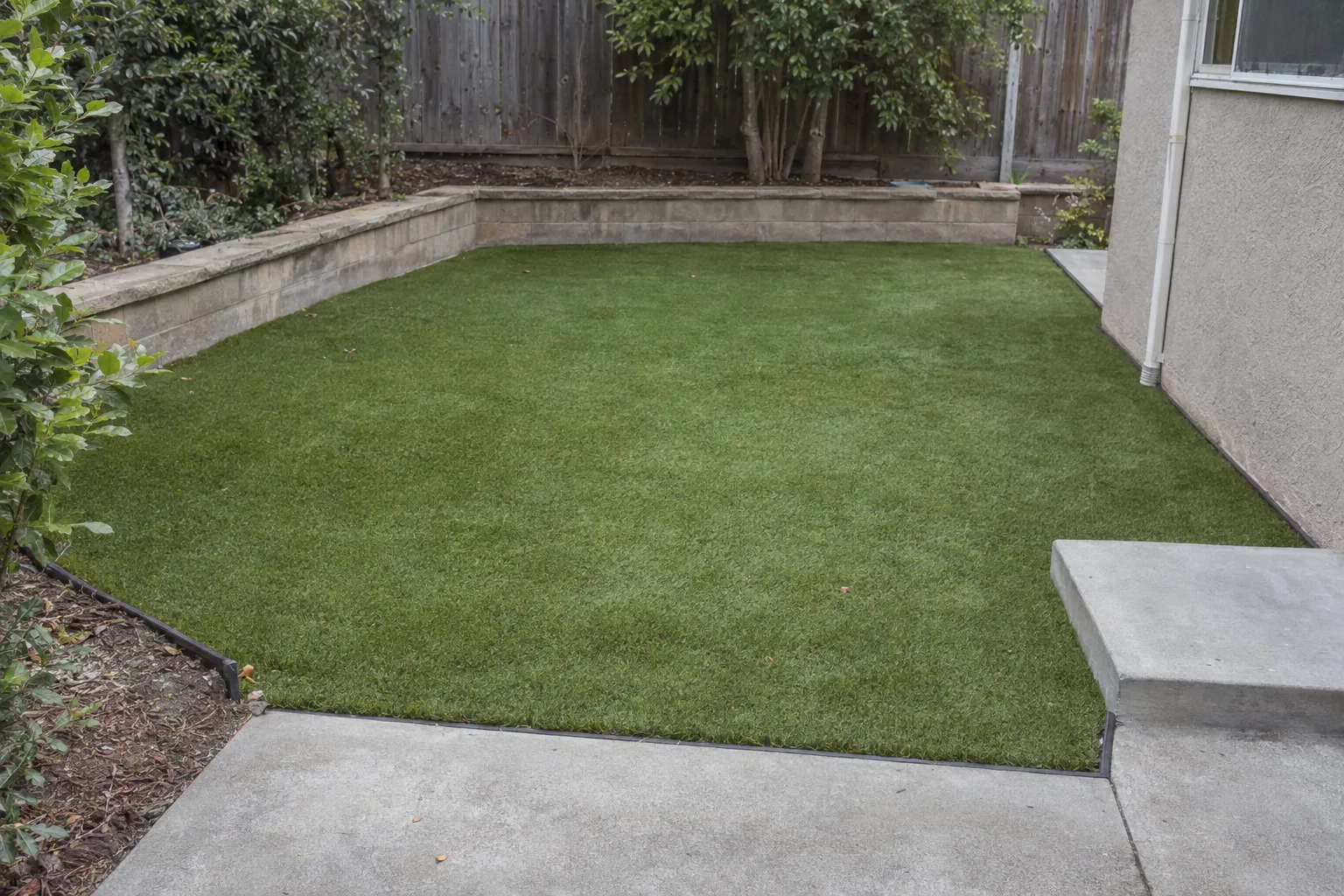 Installed artificial turf with crisp rubber-edging borders alongside a concrete step and low retaining wall in a sunny Los Angeles residential backyard, showing consistent pile texture and clean perimeter