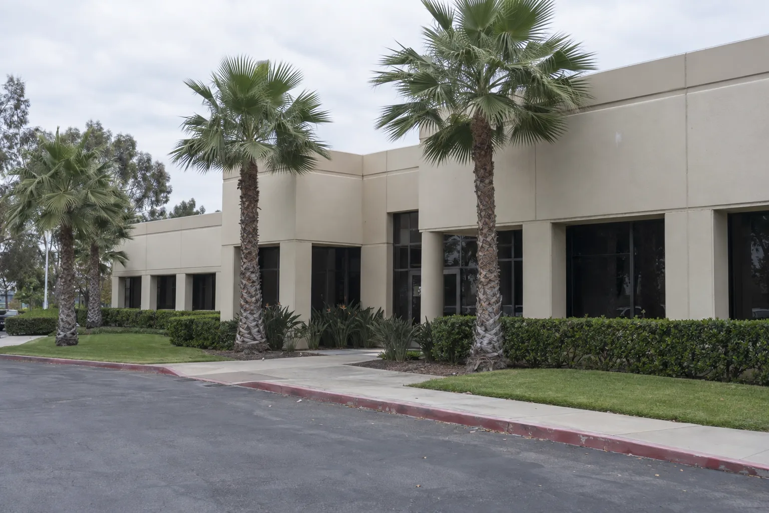 Manicured commercial grounds outside a low-rise stucco Los Angeles office building with edged turf, trimmed hedges, clean walkways, and Mexican fan palms lining the entry under midday sun