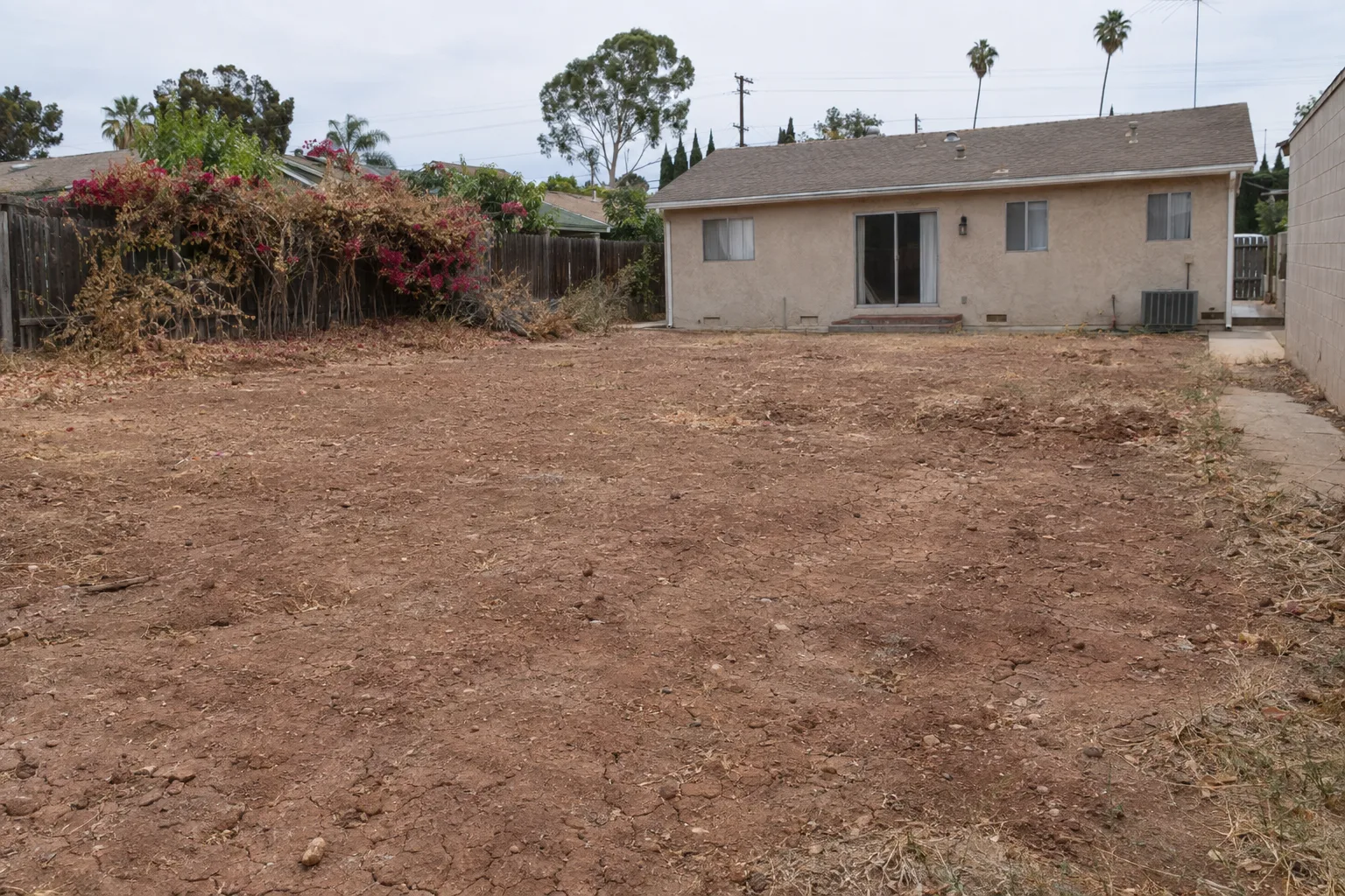 A Los Angeles backyard before sod installation, showing bare compacted clay soil with cracked dry earth, a stucco house in the background, and a wood fence along the perimeter
