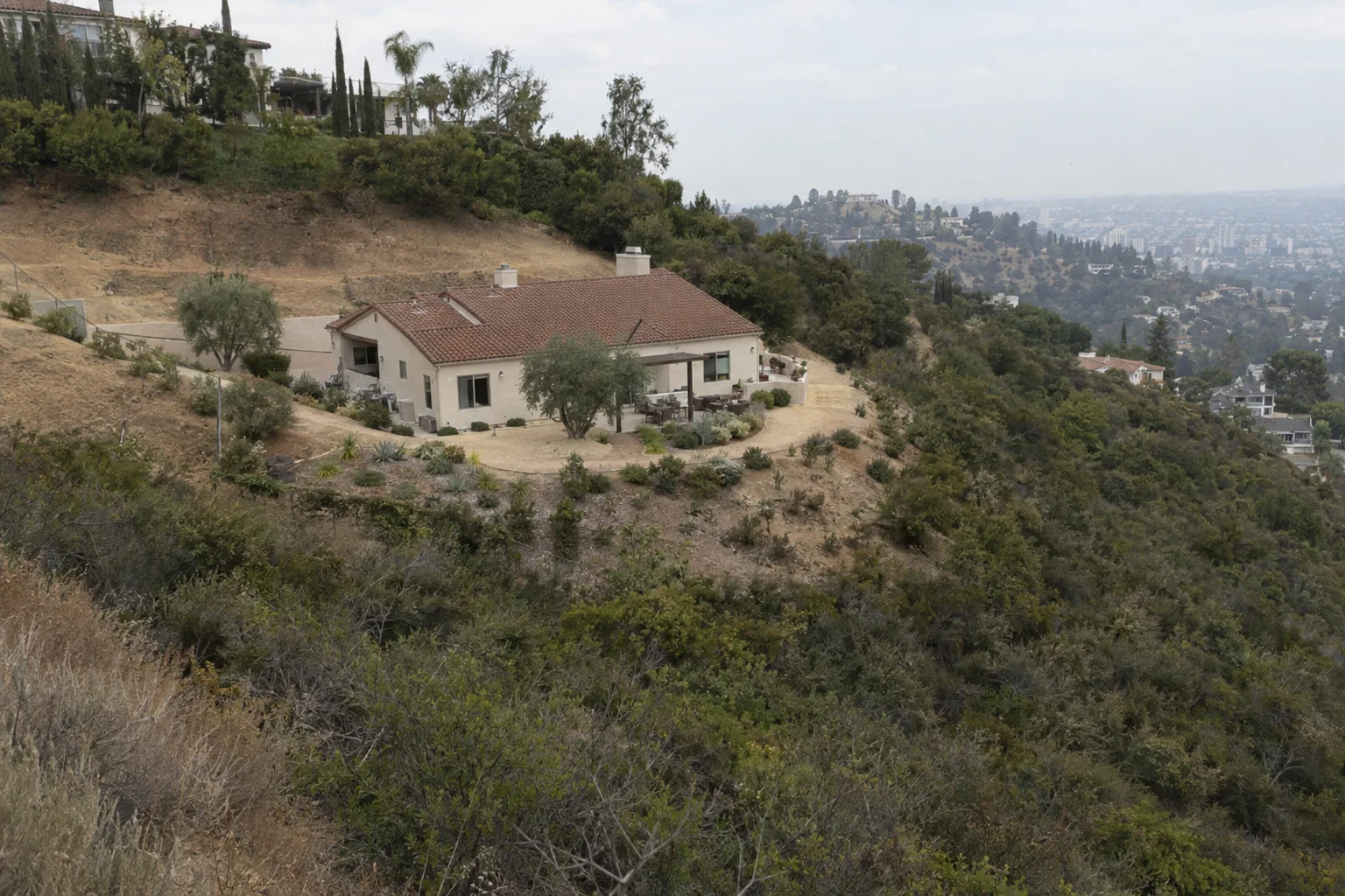 Aerial view of a hillside Los Angeles home surrounded by tiered defensible space clearance zones, with dry brush cleared near the house, sparse native plants in the middle zone, and dense chaparral in the distance