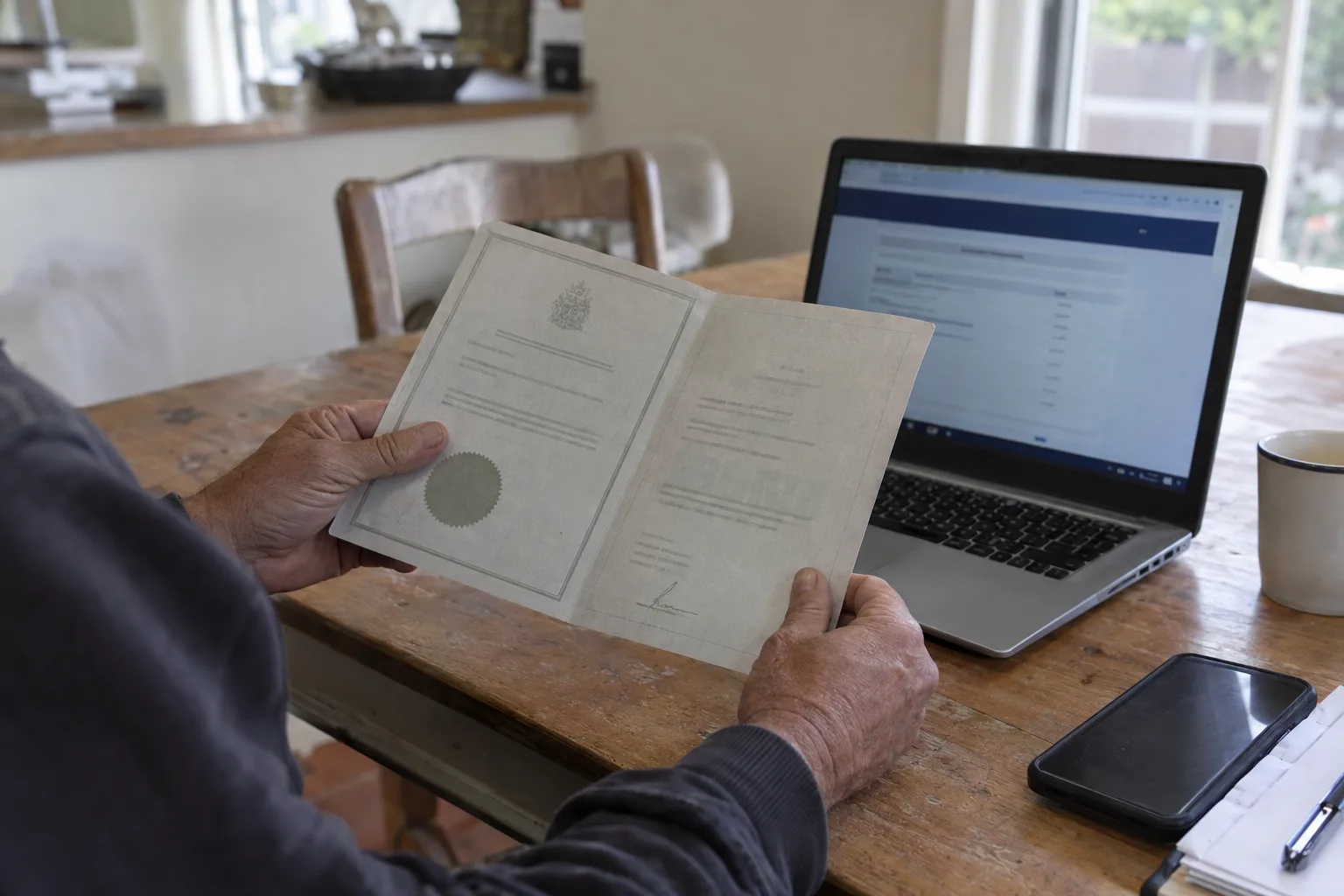 Weathered hands holding a printed contractor document beside an open laptop on a kitchen table, no legible text visible, in soft natural window light