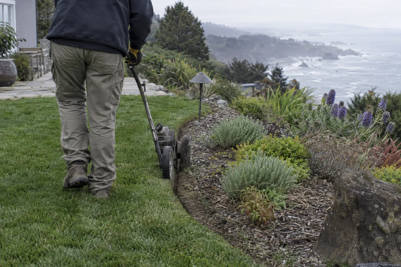 A landscaper edging the curved border between a green lawn and a planted garden bed on a sloped Palos Verdes property, with a hazy Pacific Ocean coastline visible in the background on an overcast morning.