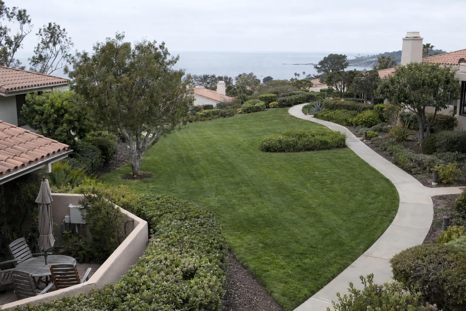 Aerial view of a Palos Verdes HOA common area featuring evenly mowed turf, trimmed shrub borders, and a curved concrete walking path, with tile-roofed residential homes visible at the edges under overcast coastal light.